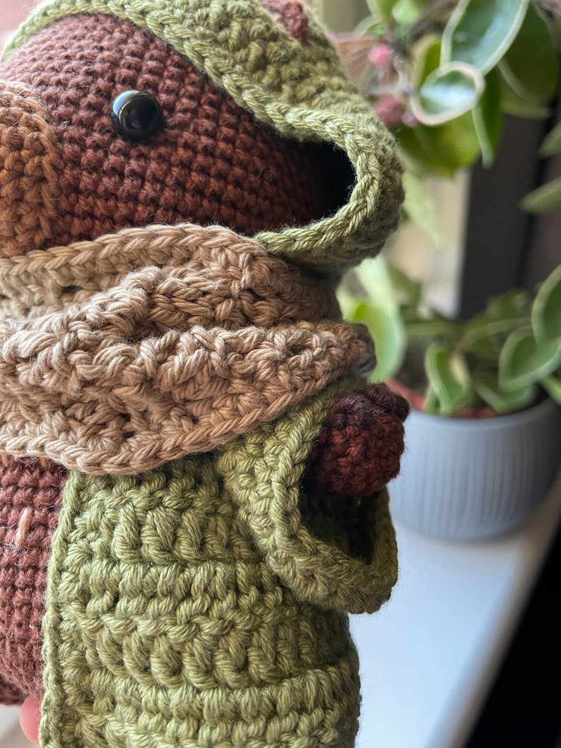 May include: A crocheted brown bear wearing a green hooded scarf and a tan scarf. The bear has black eyes and a brown nose.