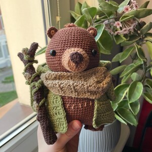 May include: A handmade crochet bear doll with a brown body, tan snout, and black eyes. The bear wears a green and tan scarf and jacket, and holds a brown stick. The bear is a whimsical, handcrafted item.