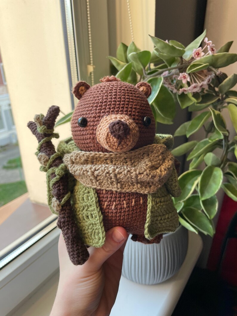 May include: A handmade crochet bear doll with a brown body, tan snout, and black eyes. The bear wears a green and tan scarf and jacket, and holds a brown stick. The bear is a whimsical, handcrafted item.
