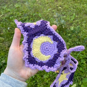 May include: A crocheted headband featuring a purple, yellow and white colour scheme. The headband has a circular design with a crescent moon shape in the centre.