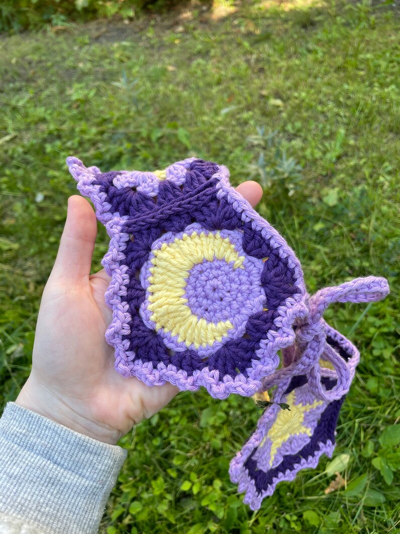 May include: A crocheted headband featuring a purple, yellow and white colour scheme. The headband has a circular design with a crescent moon shape in the centre.