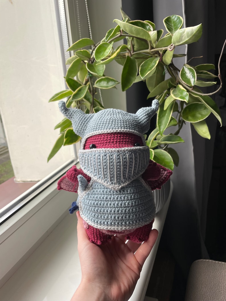 Puede incluir: Un juguete de peluche de drag&oacute;n tejido a crochet con una armadura de caballero gris y blanca. El drag&oacute;n es rojo y tiene una peque&ntilde;a cola azul.