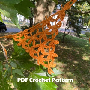 Op de afbeelding: Een close-up van een oranje haakpatroon met een bladontwerp. Het haakwerk hangt aan een tak. De tekst "PDF Crochet Pattern" is zichtbaar onderaan de afbeelding.