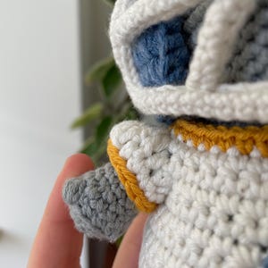May include: A crocheted plush toy with a white body, blue head, and yellow trim. The toy has a gray arm and a white helmet.