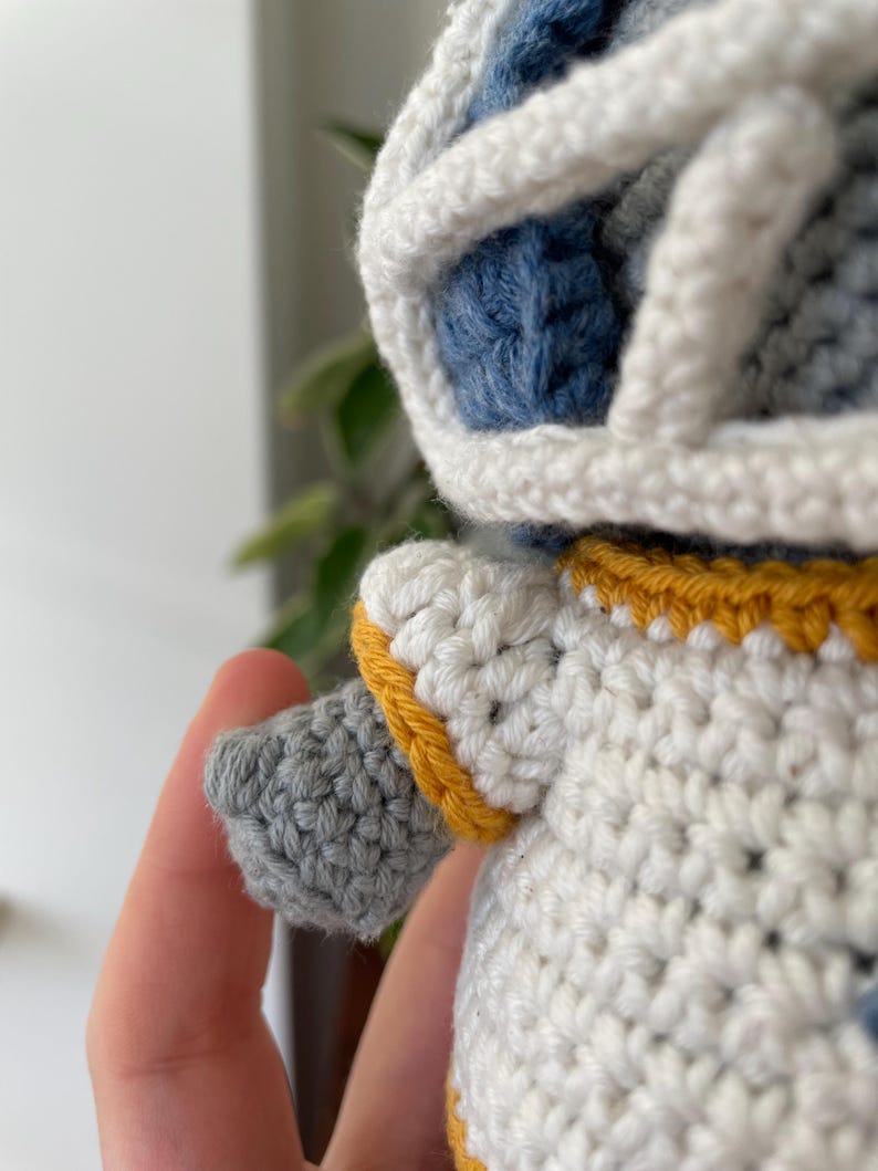 May include: A crocheted plush toy with a white body, blue head, and yellow trim. The toy has a gray arm and a white helmet.