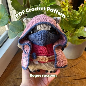 May include: Crochet pattern for a rogue raccoon wearing a pink hooded cloak, a red shirt, and a blue mask. The pattern is available as a PDF download.  "Rogue raccoon" is written below the image.