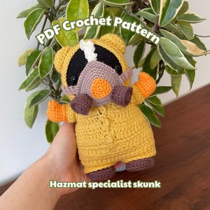 Puede incluir: Un zorrillo de ganchillo hecho a mano con un traje de protección amarillo, con guantes naranjas y un respirador morado. El zorrillo tiene una cabeza en blanco y negro. El texto de la imagen dice "PDF Crochet Pattern" y "Hazmat specialist skunk".
