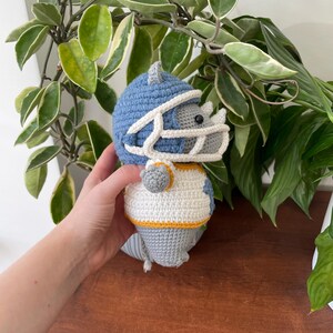 May include: A gray crocheted rhinoceros wearing a blue and white football helmet and jersey. The rhinoceros is holding a football.