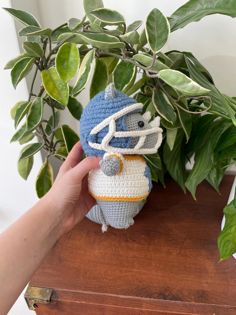 May include: A gray crocheted rhinoceros wearing a blue and white football helmet and jersey. The rhinoceros is holding a football.