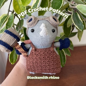 Pode incluir: Um brinquedo de rinoceronte de crochê vestindo um macacão marrom e segurando um martelo. O brinquedo tem um corpo cinza, olhos pretos e um chifre branco. O texto "Blacksmith rhino" está escrito abaixo do brinquedo.