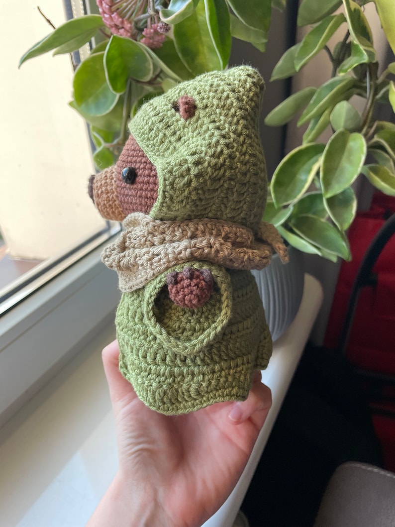 May include: A crocheted stuffed toy with a green hooded cloak and brown face. The toy is holding its hand up.