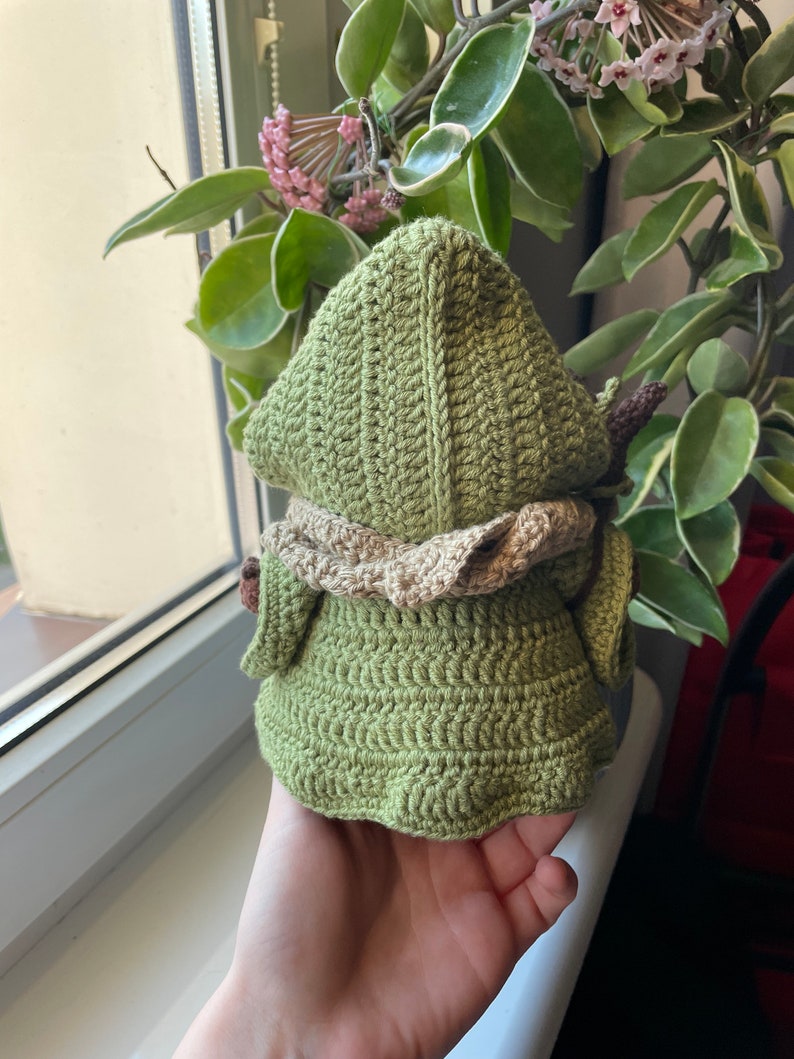May include: Green crocheted amigurumi doll with a brown crocheted hood and a brown crocheted collar. The doll is wearing a green crocheted dress.