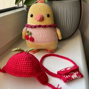 May include: A crocheted yellow chick with a pink dress and a cherry design, a red crocheted apple hat with a green leaf, and a red and white crocheted purse.
