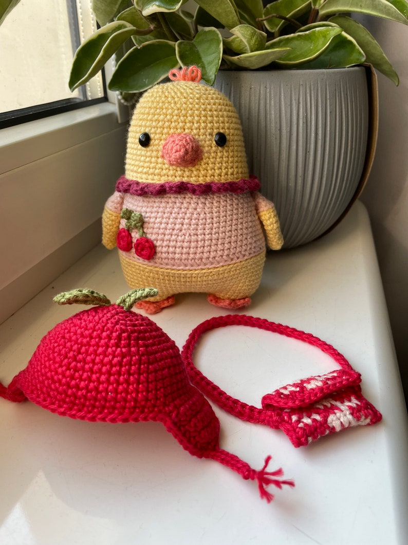 May include: A crocheted yellow chick with a pink dress and a cherry design, a red crocheted apple hat with a green leaf, and a red and white crocheted purse.