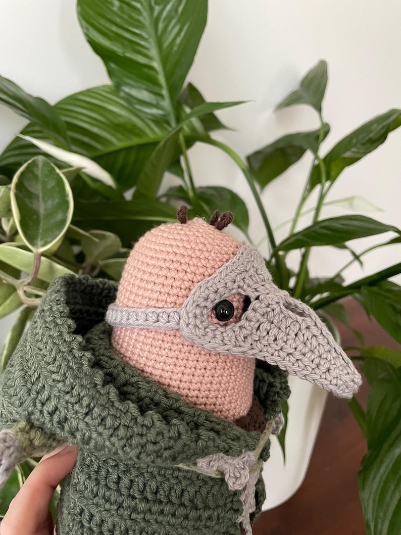 May include: A crocheted plush toy wearing a gray crocheted mask with a long beak. The toy is wearing a green crocheted hooded jacket.