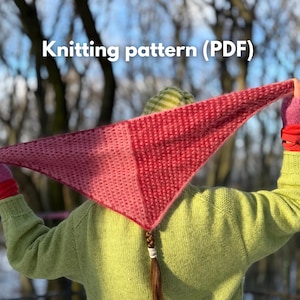 May include: A knitted triangular shawl in shades of pink and red is held up against a blurred outdoor background. The shawl is made with a textured stitch pattern. The text "Knitting pattern (PDF)" is visible at the top of the image.