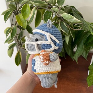 May include: A crocheted gray rhinoceros wearing a blue and white football helmet and jersey, holding a brown and orange football.