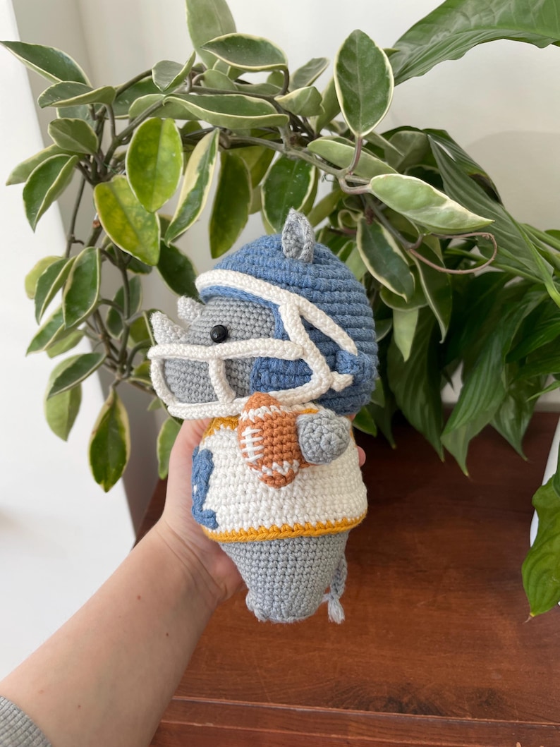 May include: A crocheted gray rhinoceros wearing a blue and white football helmet and jersey, holding a brown and orange football.