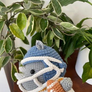 May include: A gray crocheted rhinoceros wearing a blue and white football helmet and jersey, holding a brown football. The rhinoceros is holding the football in its right hand.