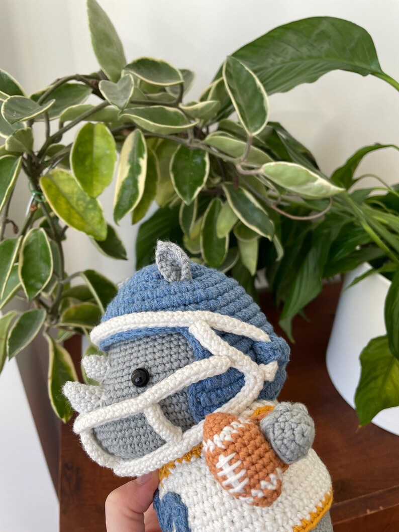 May include: A gray crocheted rhinoceros wearing a blue and white football helmet and jersey, holding a brown football. The rhinoceros is holding the football in its right hand.