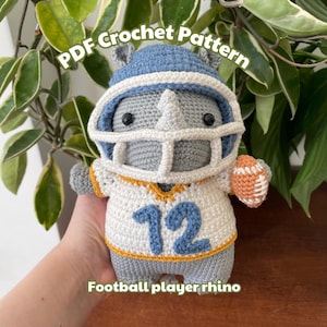 May include: Crocheted gray rhino wearing a blue and white football helmet and jersey with the number 12. The rhino is holding a brown and orange football. The text "PDF Crochet Pattern" is above the rhino and the text "Football player rhino" is below the rhino.