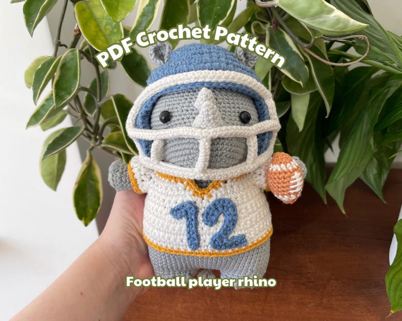 May include: Crocheted gray rhino wearing a blue and white football helmet and jersey with the number 12. The rhino is holding a brown and orange football. The text "PDF Crochet Pattern" is above the rhino and the text "Football player rhino" is below the rhino.