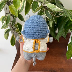 May include: A crocheted gray hippopotamus wearing a white and yellow crocheted sweater. The hippopotamus is holding a small crocheted object in its right hand.