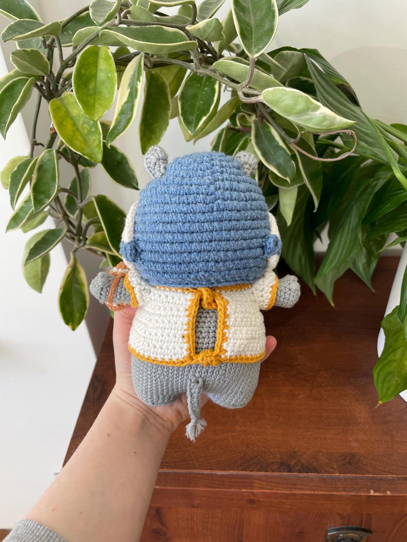 May include: A crocheted gray hippopotamus wearing a white and yellow crocheted sweater. The hippopotamus is holding a small crocheted object in its right hand.