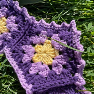 May include: A purple and yellow crocheted granny square blanket with a flower pattern. The blanket is lying on a bed of green grass.