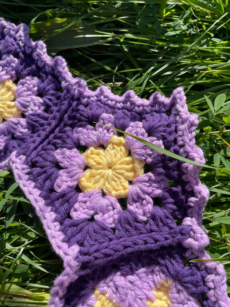May include: A purple and yellow crocheted granny square blanket with a flower pattern. The blanket is lying on a bed of green grass.