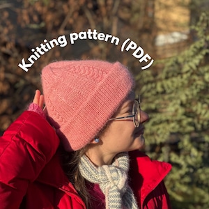 Op de afbeelding: Een roze gebreide muts met een textuurontwerp wordt getoond. De muts wordt gedragen door een persoon die een rode jas en een sjaal met patroon draagt. De tekst "Knitting pattern (PDF)" wordt boven de muts weergegeven.