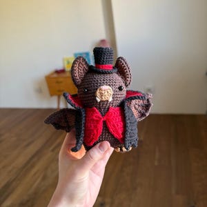Può includere: Bambola a forma di pipistrello all'uncinetto con cappello a cilindro nero con fascia rossa, papillon rosso e mantello nero e rosso. Il pipistrello ha pelliccia marrone, piccoli occhi neri e muso beige. Le ali sono marrone scuro.