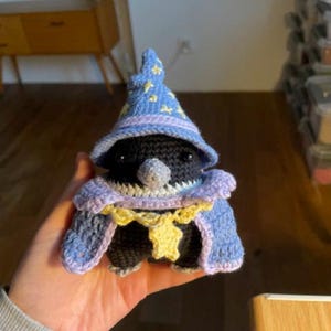 Puede incluir: Un peluche de ganchillo con forma de pájaro negro que lleva un sombrero de mago azul con patrones de estrellas amarillas, un cuello de color violeta claro y una capa azul a juego con un colgante amarillo en forma de estrella en el frente.