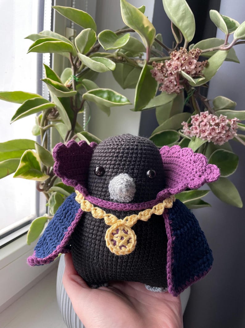 May include: A black crocheted bird wearing a purple cape and a gold chain with a yellow pendant. The bird has black eyes and a gray beak.