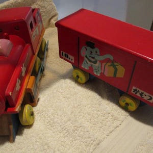 May include: A red wooden toy train with a caboose. The train has a black and blue wheel and a white elephant with a red gift box on the side. The caboose has the Japanese word "かぎつ" written on the side.