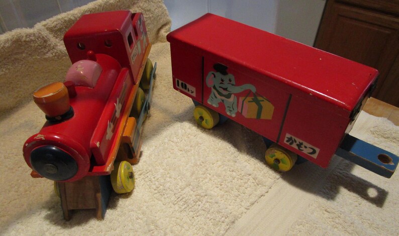 May include: A red wooden toy train with a caboose. The train has a black and blue wheel and a white elephant with a red gift box on the side. The caboose has the Japanese word "かぎつ" written on the side.
