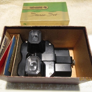 Peut inclure: Un ensemble stéréo View-Master vintage dans une boîte brune avec un visionneur noir et argent et une pile de bobines View-Master colorées. La boîte porte l'inscription "View-Master Stereo Set".