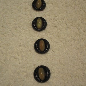 May include: Four black buttons with oval, light brown centers. Each button has a dark outer ring with a subtle, ridged design. The buttons are arranged vertically on a textured, off-white surface.