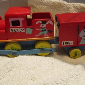 May include: A red wooden toy train with a blue and yellow engine and a red boxcar. The engine has a white rabbit with a blue and white scarf and a green flag. The boxcar has a blue elephant with a red hat and a yellow box with a red bow. The train has the Japanese text "まかいで" and "かぞく" on it.