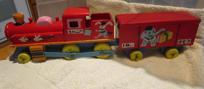 May include: A red wooden toy train with a blue and yellow engine and a red boxcar. The engine has a white rabbit with a blue and white scarf and a green flag. The boxcar has a blue elephant with a red hat and a yellow box with a red bow. The train has the Japanese text "まかいで" and "かぞく" on it.