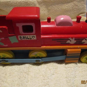 May include: A red wooden toy train with a white rabbit and a blue flag on the side. The train has a pink caboose and a brown chimney. The train says "さかんじゃ" on the side.