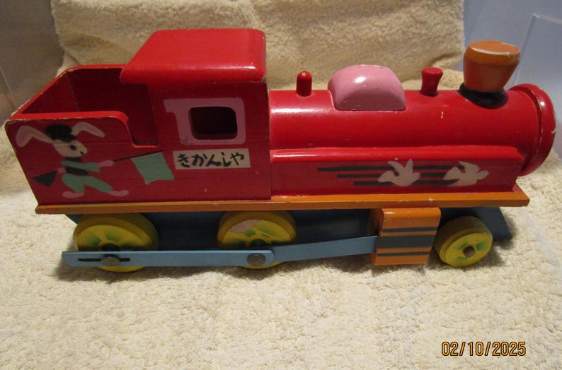 May include: A red wooden toy train with a white rabbit and a blue flag on the side. The train has a pink caboose and a brown chimney. The train says "さかんじゃ" on the side.