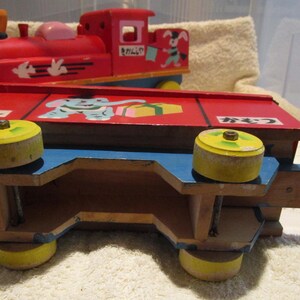 May include: A red wooden toy train with a blue and yellow caboose. The train has a painted elephant, a gift box, and the Japanese word "プレゼント" on the side. The train has wheels with yellow rims.