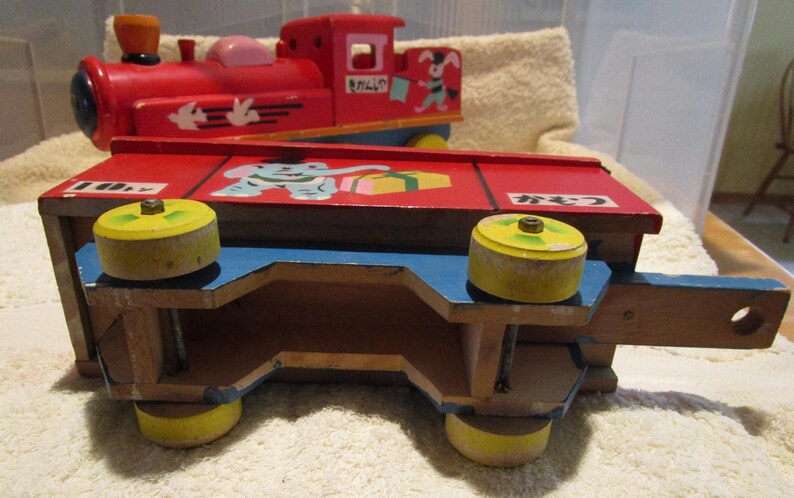 May include: A red wooden toy train with a blue and yellow caboose. The train has a painted elephant, a gift box, and the Japanese word "プレゼント" on the side. The train has wheels with yellow rims.