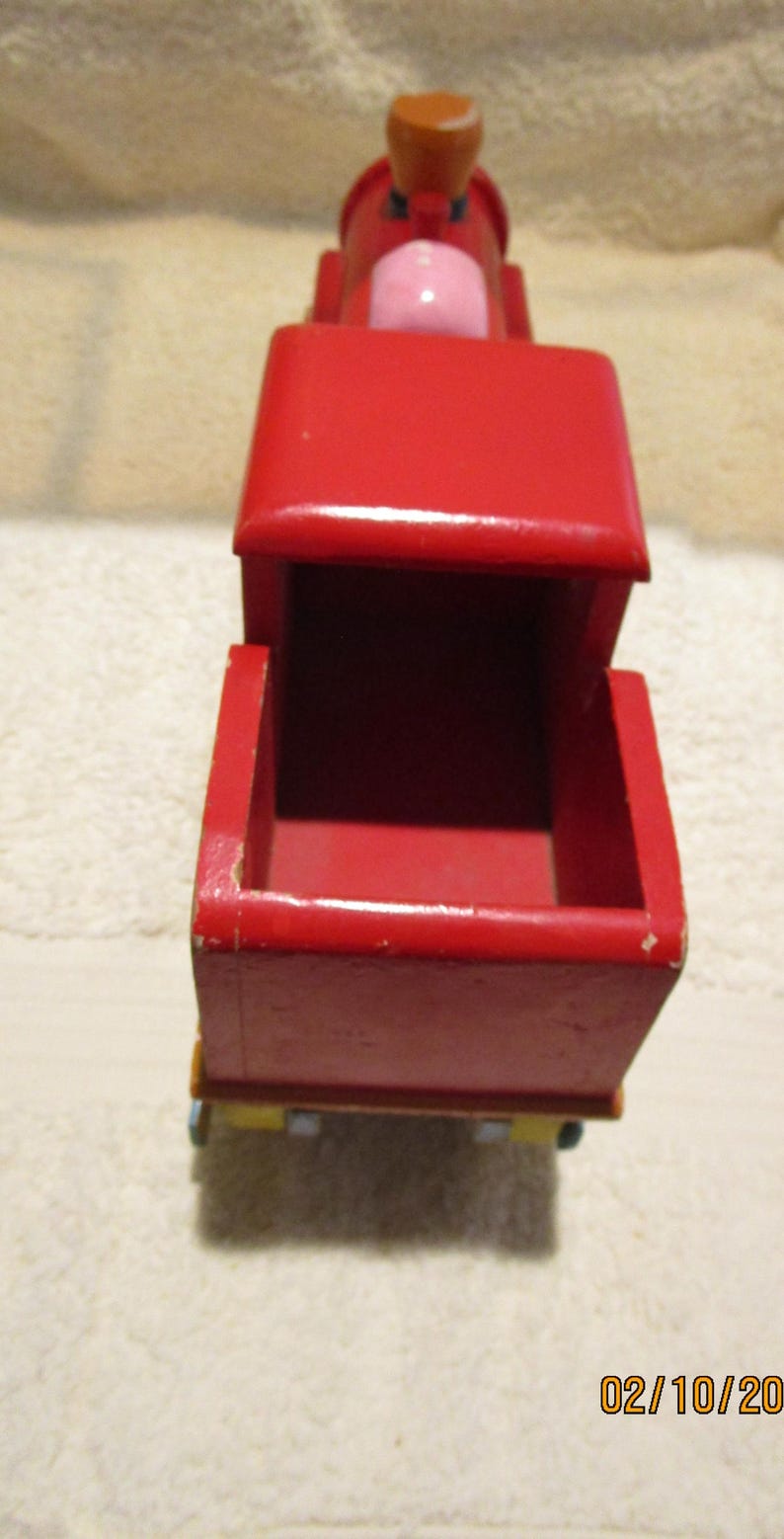 May include: A red wooden toy train engine with a pink and brown cab. The engine has a removable top and a yellow and green base.