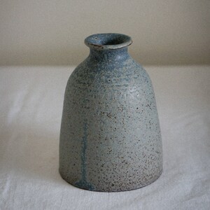 Op de afbeelding: Een kleine, lichtblauwe keramische vaas met een gespikkeld glazuur. De vaas heeft een rond lichaam en een smalle opening.