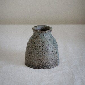 Op de afbeelding: Een kleine, grijze keramische vaas met een gespikkeld glazuur. De vaas heeft een ronde bodem en een licht uitlopende opening.