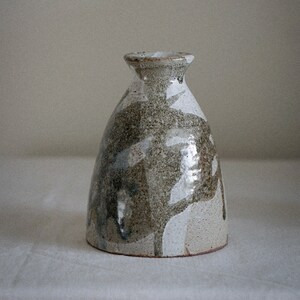 Op de afbeelding: Een kleine, witte en bruine keramische vaas met een gespikkeld glazuur. De vaas heeft een brede basis en een smalle hals.