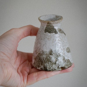 Op de afbeelding: Een kleine, witte keramische vaas met bruine en groene gespikkelde glazuur. De vaas heeft een ronde basis en een licht uitlopende opening.