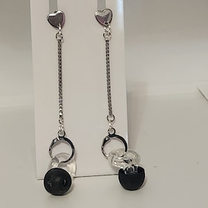 May include: A pair of dangle earrings with silver heart-shaped studs. The earrings feature a silver chain, clear circular beads, and a black, rounded bead at the bottom. The earrings are displayed on a white background.